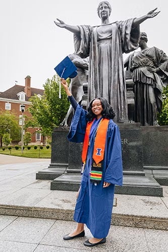 Student in graduation cap and gown by the Alma Mater