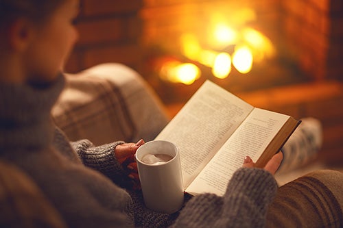 A woman sits with a book in front of a fireplace