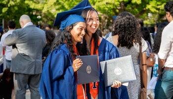 Two graduates posing for a photo in their caps and gowns, holding their diploma holders