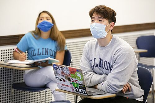 Two students listen during a class. They are both masked.