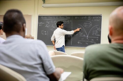 A student points to a blackboard with formulas written on it in chalk