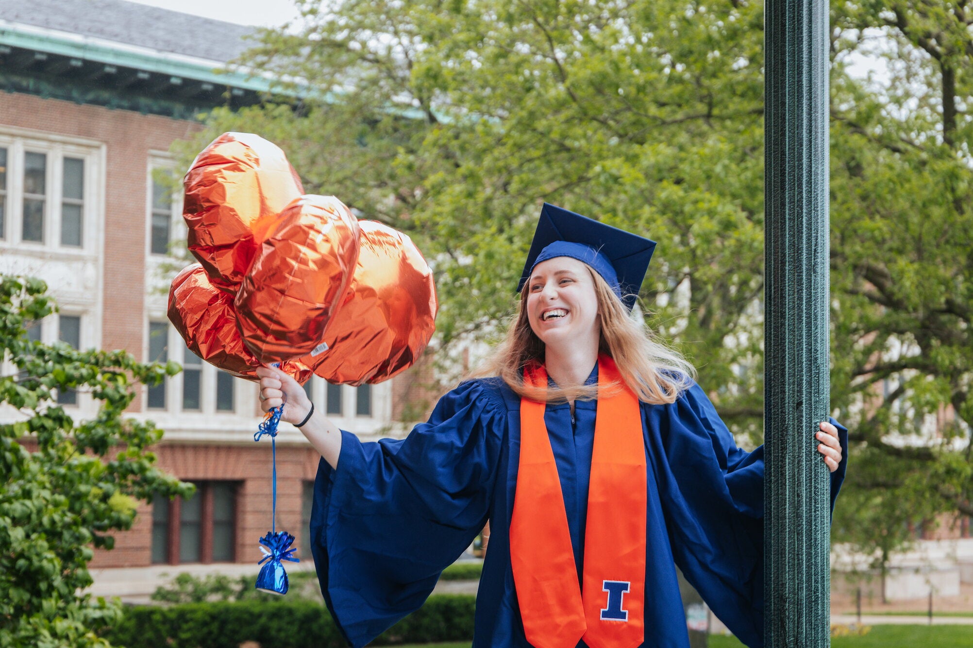Alumna in cap and gown on the Quad side of Foellinger Auditorium facing Lincoln Hall holding orange balloons.