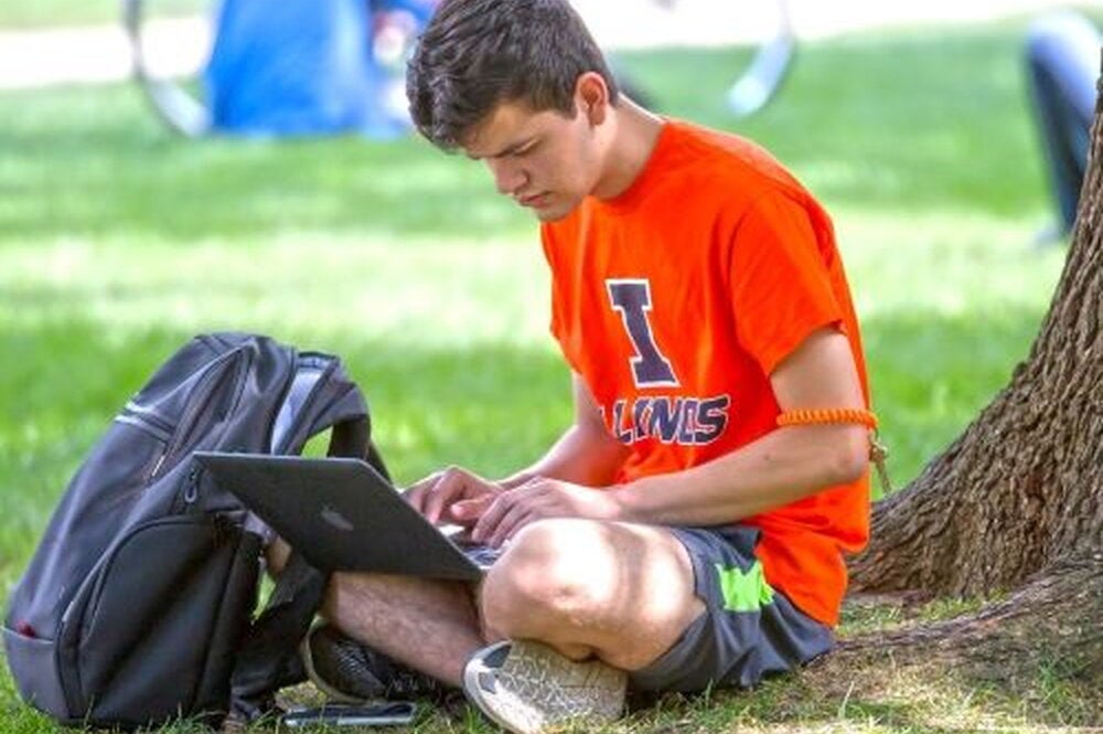 Student working on computer sitting under a tree on the Quad