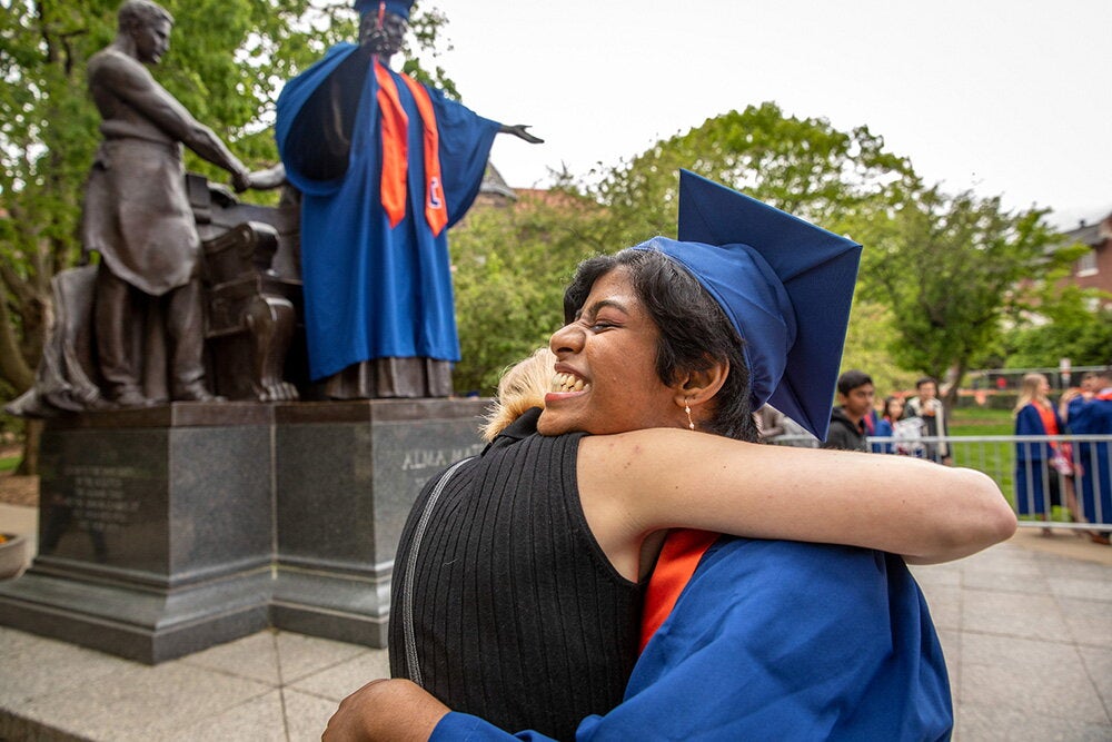 A graduate hugging a friend by the Alma Mater