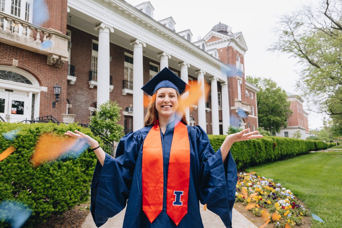 Illinois graduate in blue cap and gown, orange stole, throwing orange and blue confetti in front of the camera