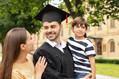 Man wearing a cap and gown holds his young son and smile as his wife
