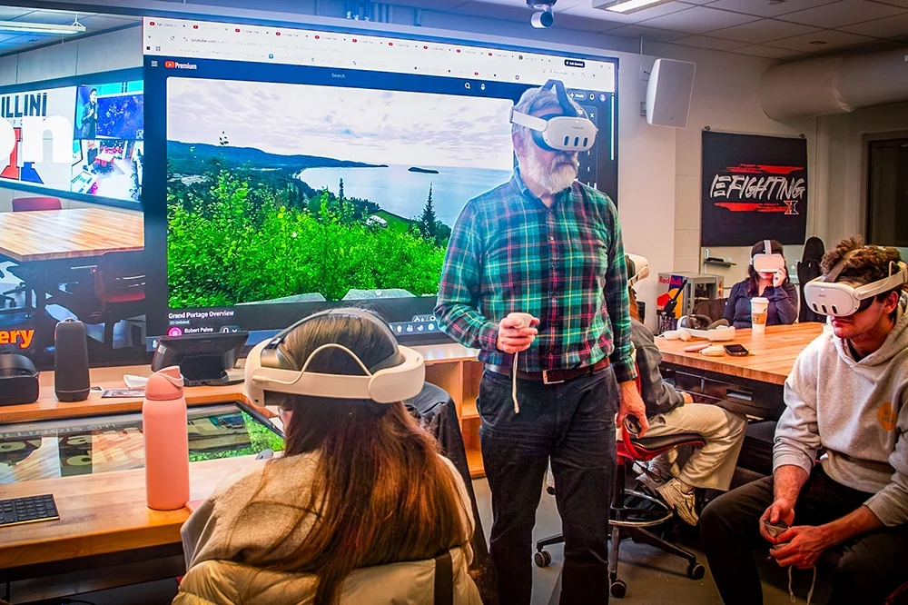Robert Pahre wearing virtual reality goggles in front of a screen showing wilderness